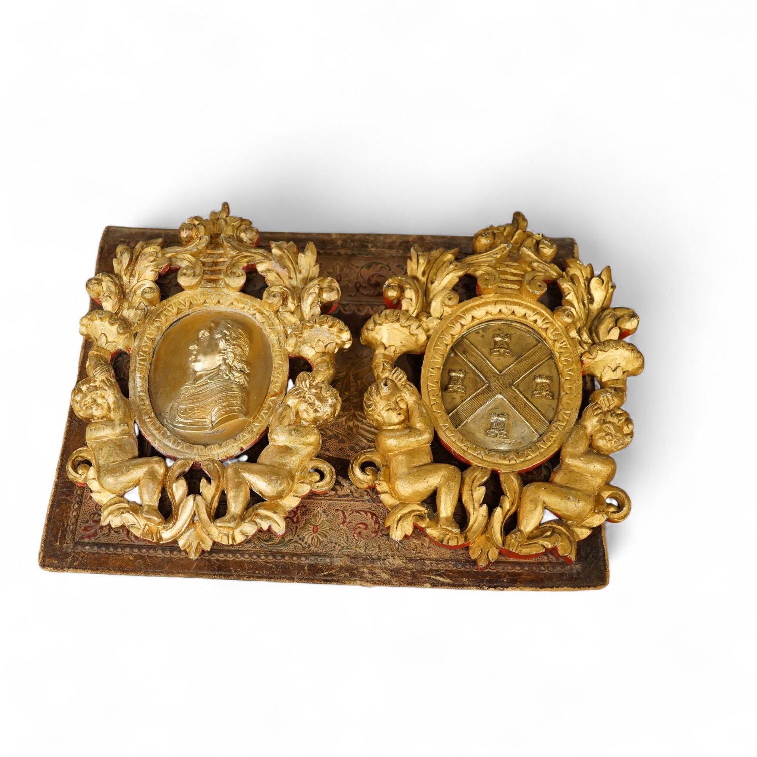 Two gilt metal plaques housed in giltwood ‘cherub’ frames and a heraldic leather folder with watered silk lining, 34 x 27cm